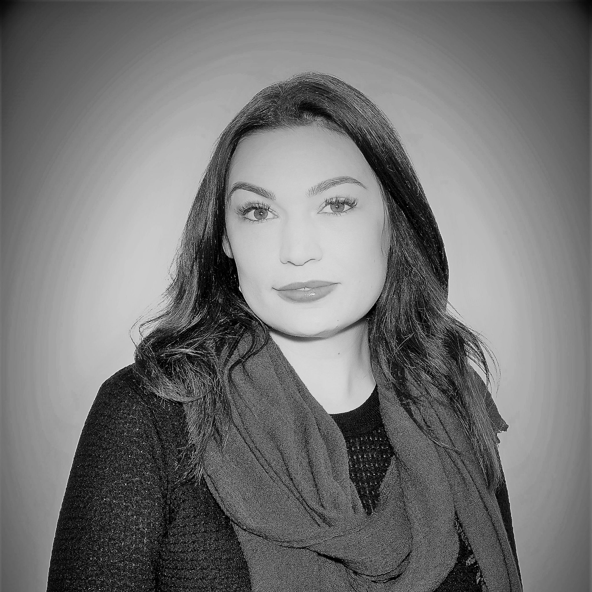 Black and white professional headshot of a woman with long dark hair, wearing a textured dark sweater and a scarf, looking directly at the camera with a neutral expression.