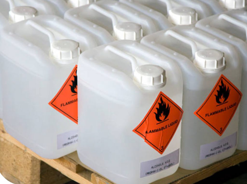 Multiple white plastic containers labeled ‘Flammable Liquid’ stored on a wooden pallet, highlighting improper or bulk storage of hazardous materials in workplace environments.